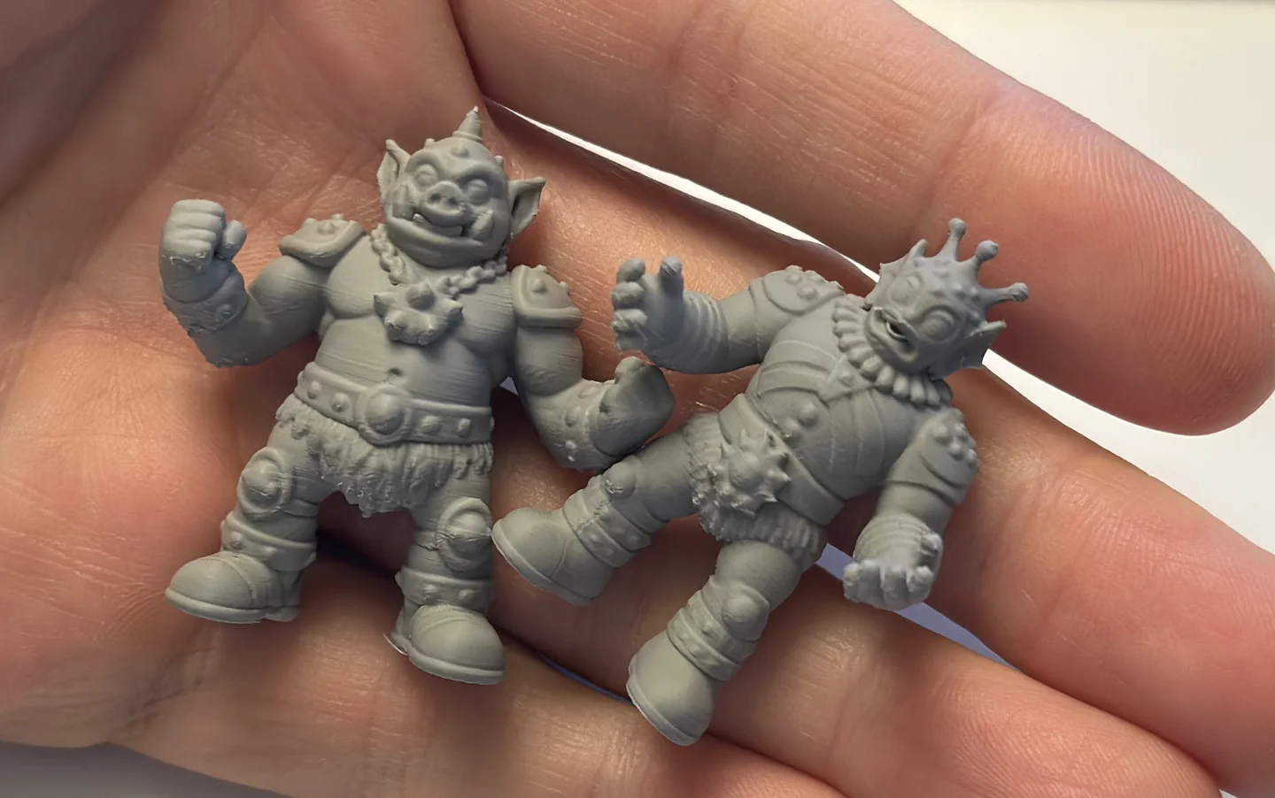 Two small gray 3D-printed figures, a boar warrior and crowned fish king, resting in Ryan’s hand.