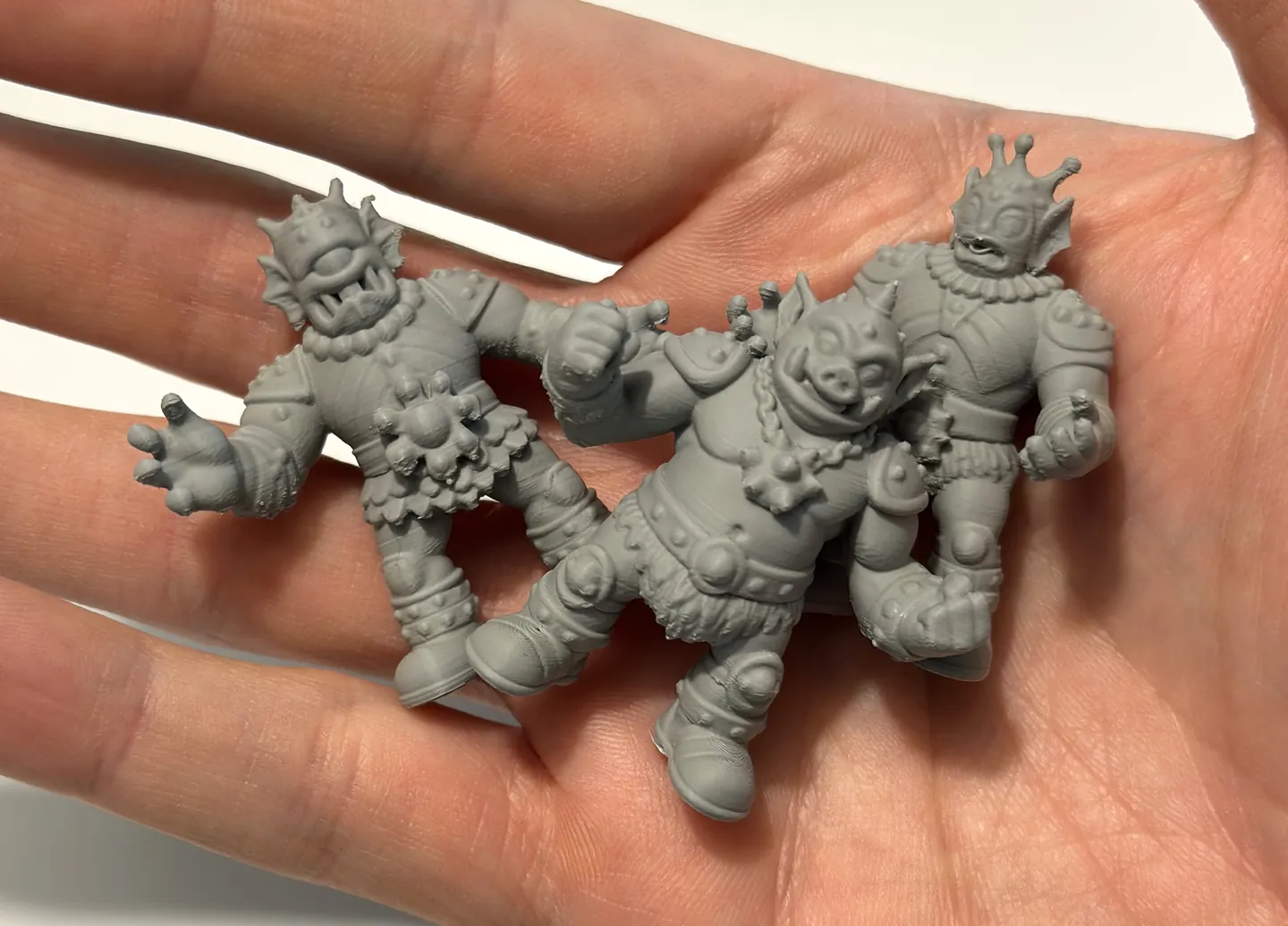 Three small gray 3D-printed figures, a cyclops warrior, a boar warrior and a crowned fish king, resting in Ryan’s open hand.